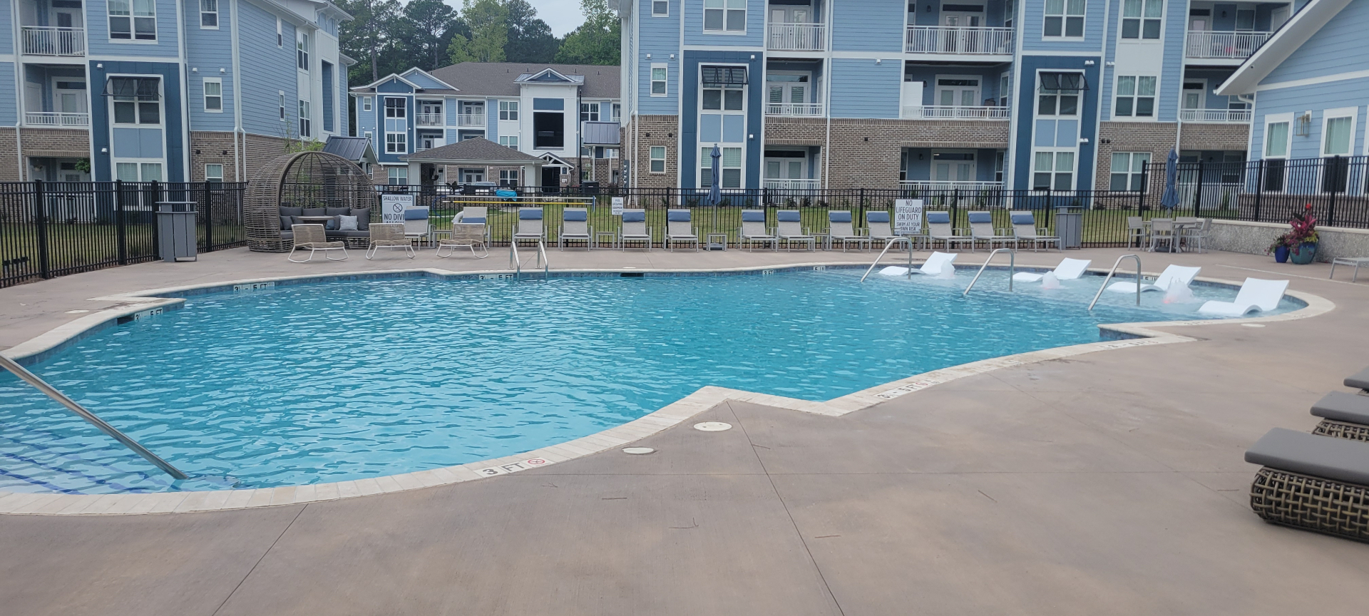Wide view of the completed pool — freeform shape, in-water loungers, and full lounge chair deck against the apartment buildings.